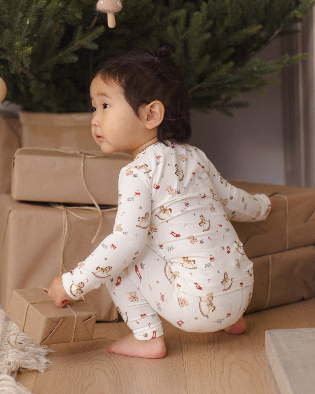 A toddler wearing a white long-sleeve pajama set with a toy chest print, crouching on a wooden floor next to wrapped gifts in front of a Christmas tree.