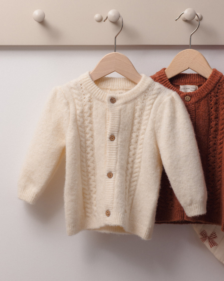 Two knitted cardigan sweaters hanging on wooden hangers against a light wall. The front sweater is cream/off-white with a fuzzy texture and brown buttons, and the sweater behind it is rust brown.