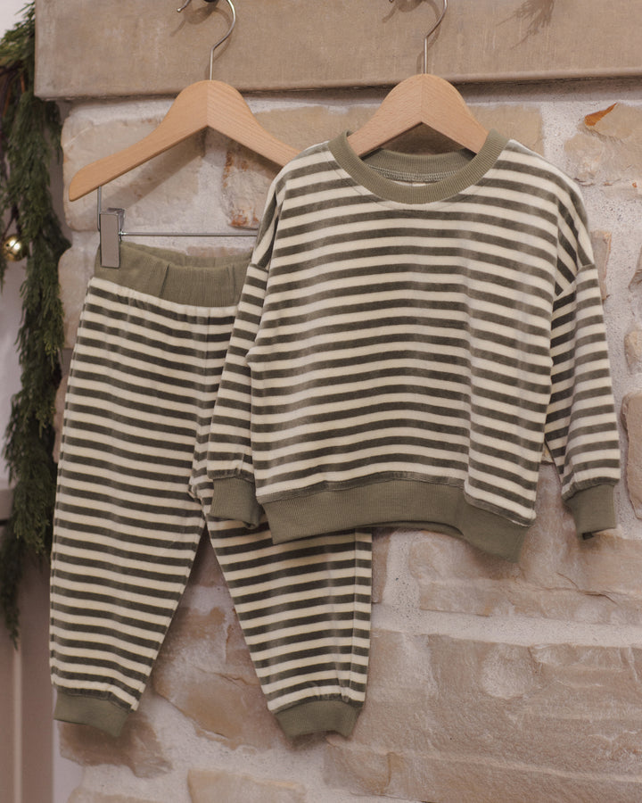 A two-piece children's pajama set with a long-sleeve top and matching pants, featuring horizontal stripes in olive green and cream. The set is displayed on wooden hangers against a rustic stone wall background.