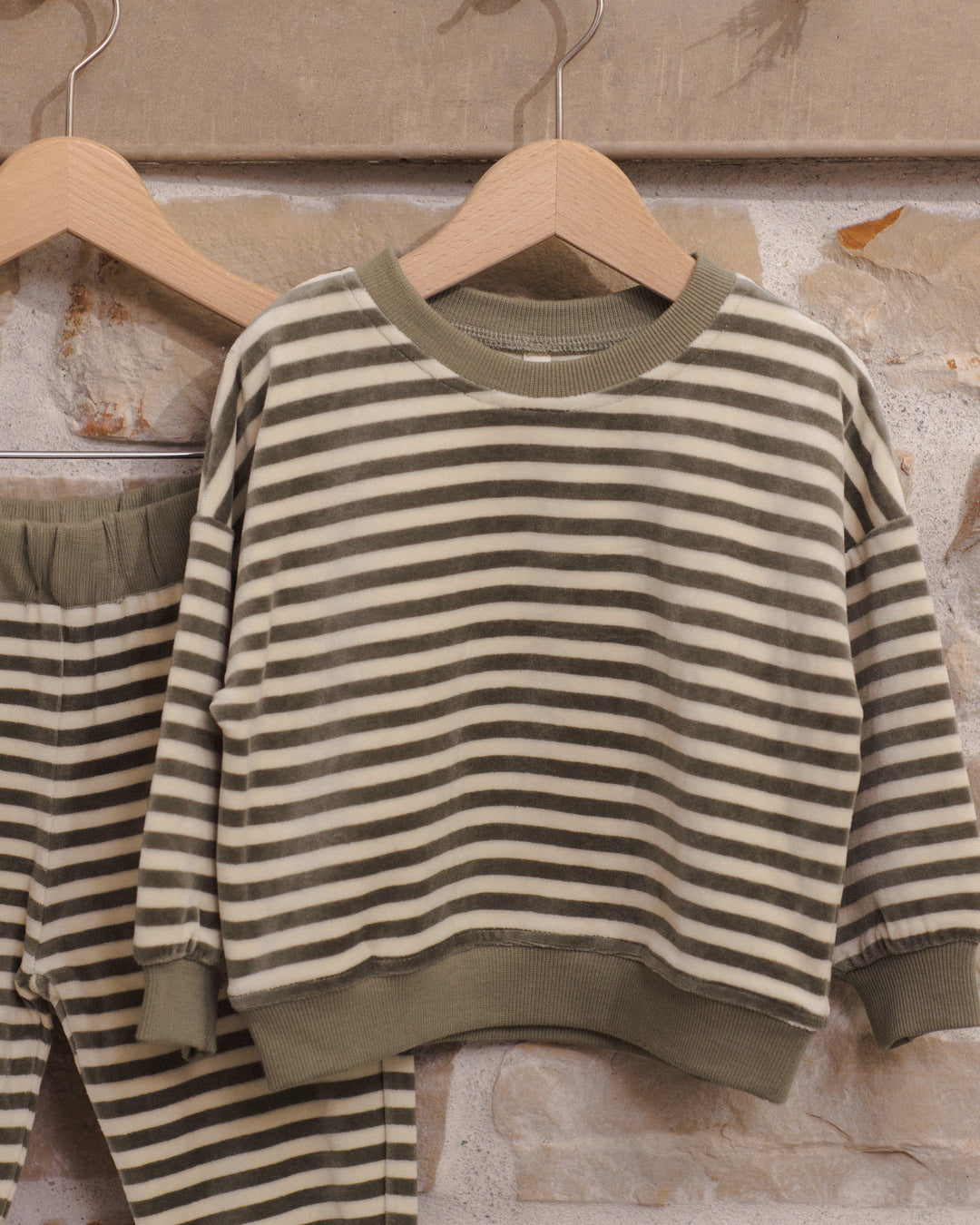 A children's two-piece lounge set featuring a long-sleeve crewneck sweatshirt and matching pants, both with horizontal olive green and cream stripes, hanging on wooden hangers against a textured stone wall.