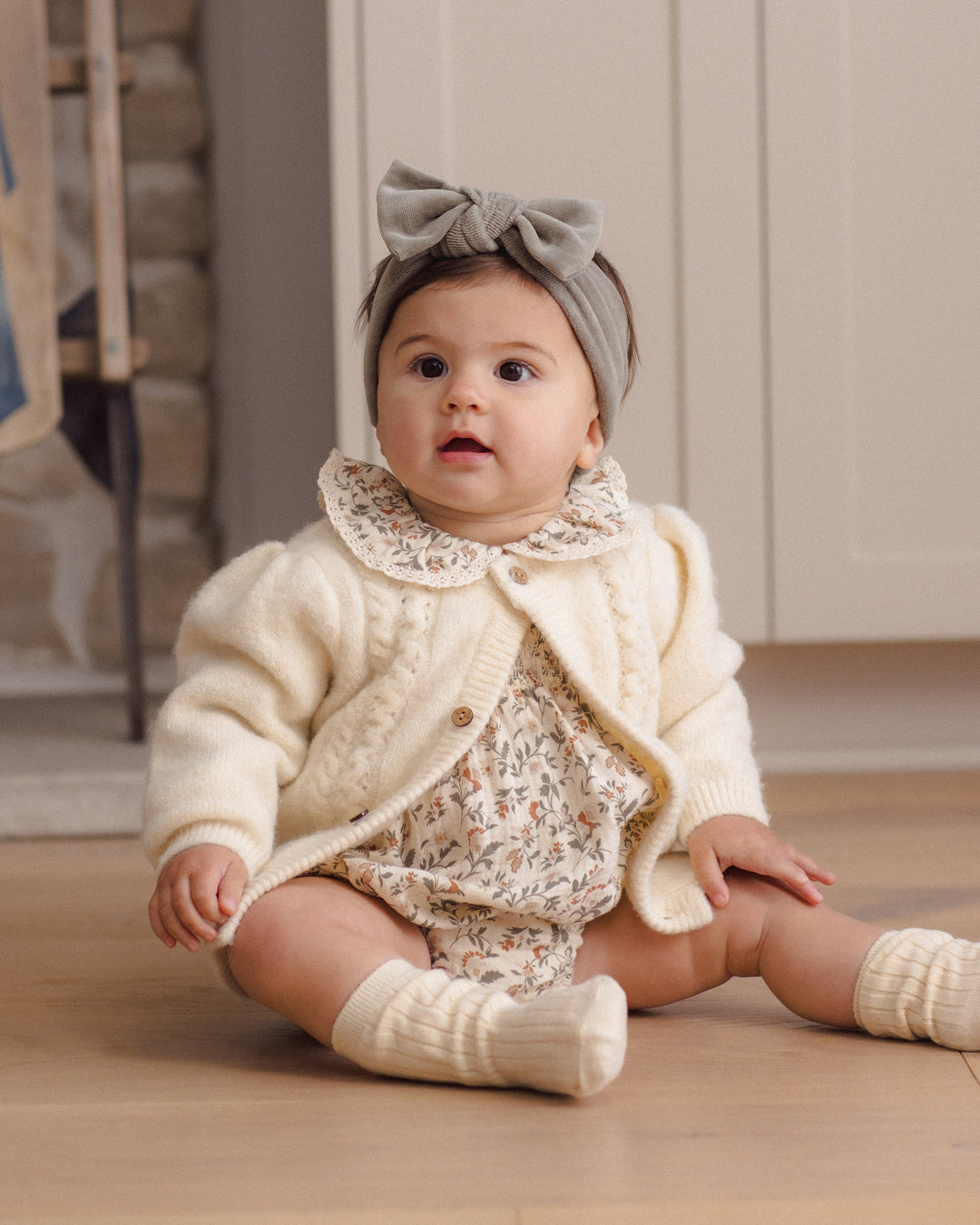 A baby sitting on a wooden floor wearing a cream cable-knit cardigan, a matching floral romper, cream knit booties, and a gray bow headband.