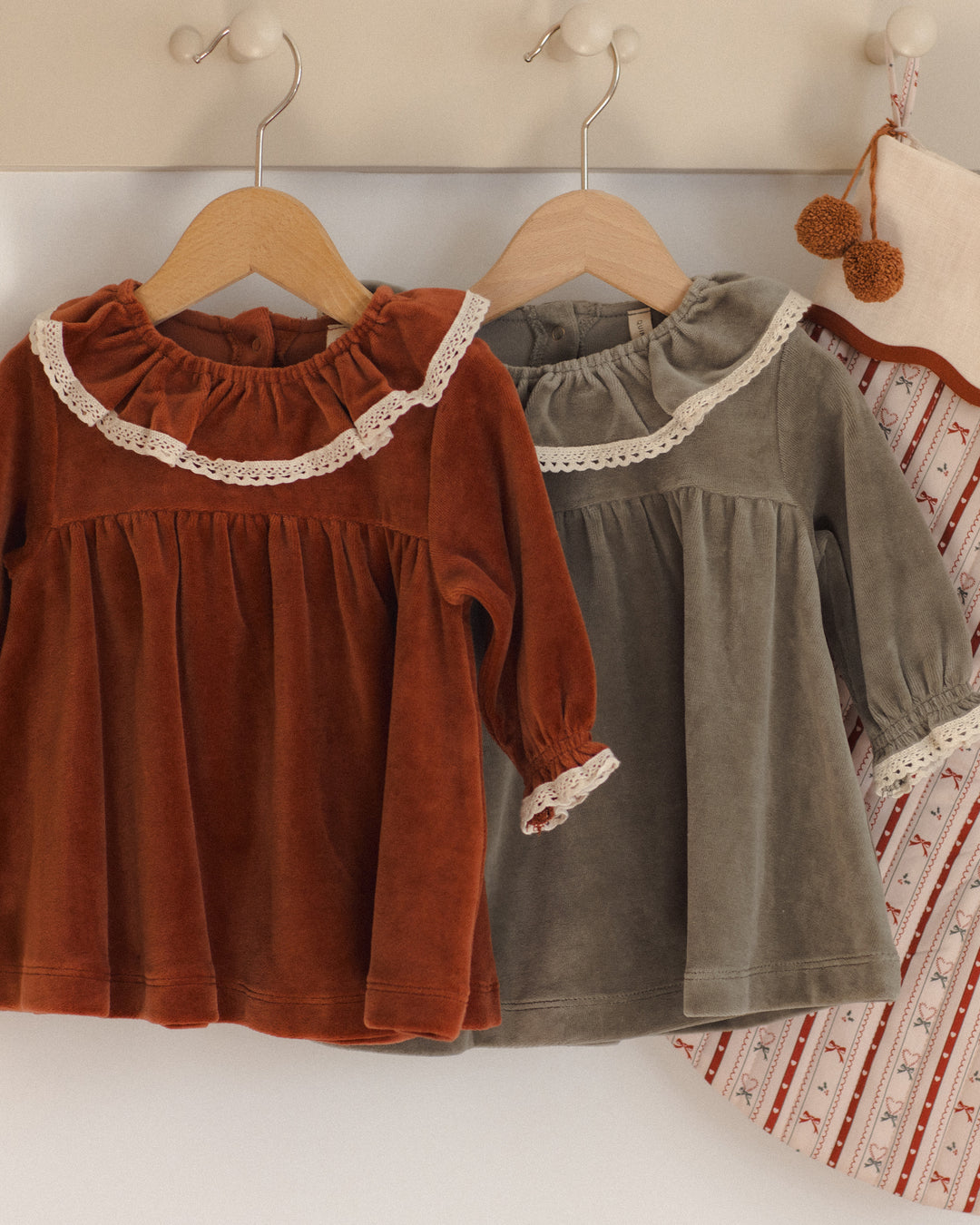 Two toddler girls' long-sleeve velvet tops hanging on wooden hangers on a wall. The front top is rust brown with a white lace ruffle collar and cuff, and the top behind it is muted gray-green with a similar white lace trim.