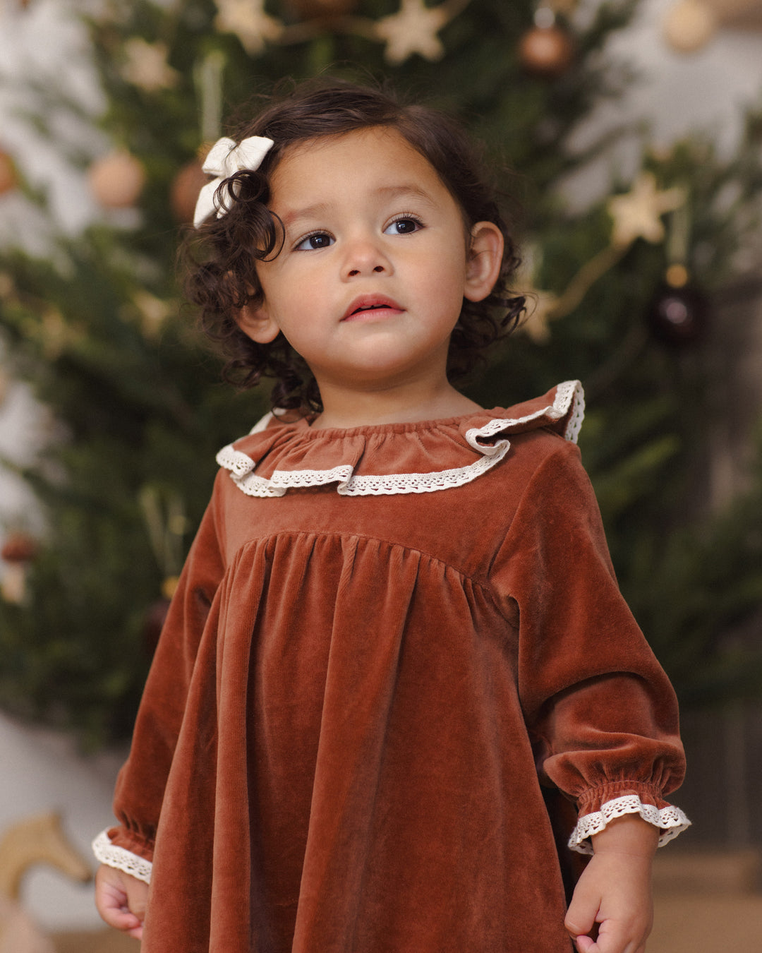 A toddler girl wearing a rust-red velvet long-sleeve dress with white lace trim, standing in front of a blurred Christmas tree.