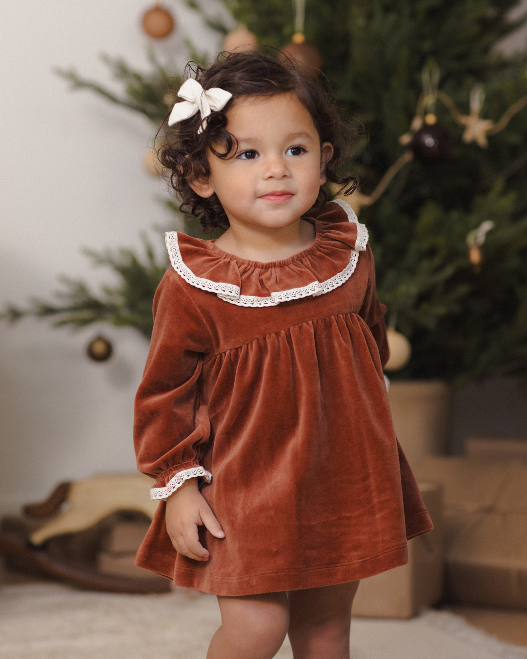 A toddler girl wearing a rust-red velvet long-sleeve dress with a white ruffled collar and cuffs, standing in front of a decorated Christmas tree.