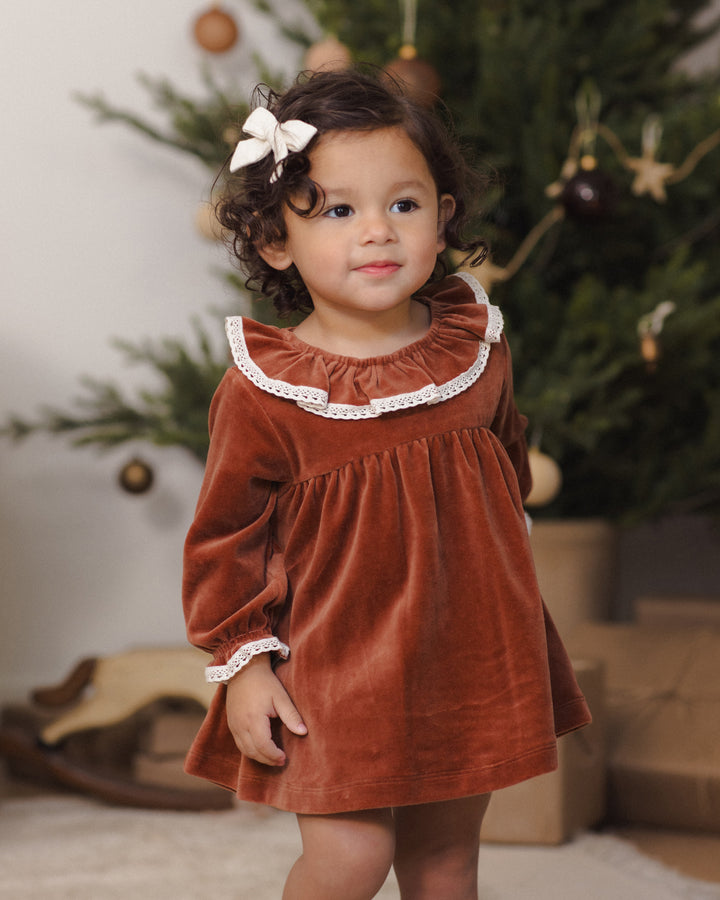 A toddler girl wearing a rust-red velvet long-sleeve dress with a white ruffled collar and cuffs, standing in front of a decorated Christmas tree.