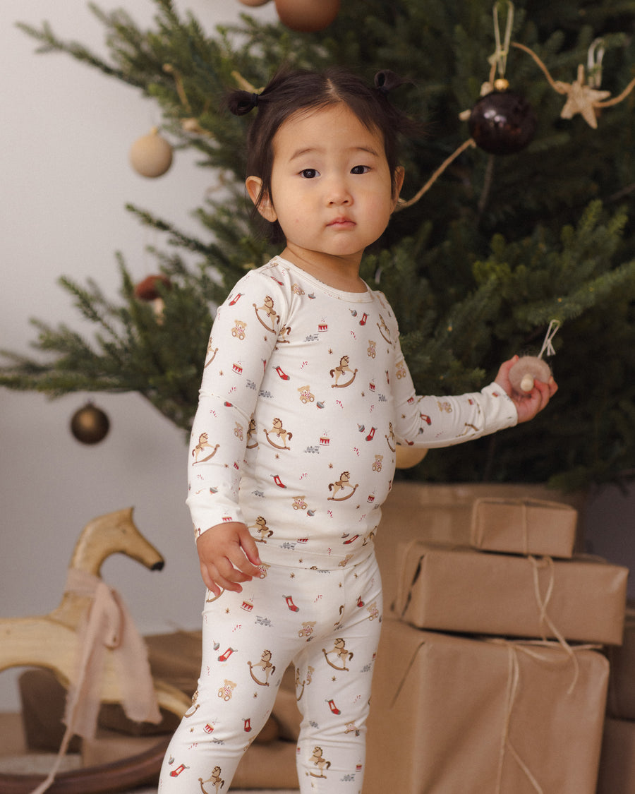 A young child wearing a white long-sleeve pajama set with a small teddy bear print stands in front of a decorated Christmas tree, holding a ornament.