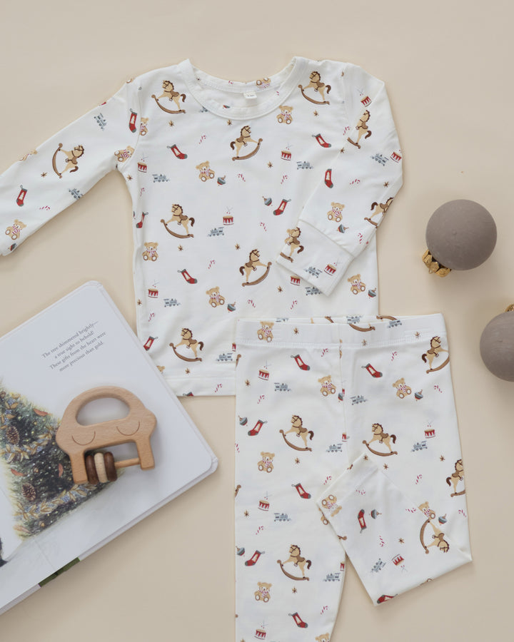 A baby long-sleeve pajama set with matching pants, featuring an all-over playful print of rocking horses, teddy bears, high heels, and small red bows on a warm cream background. The set is styled in a flat-lay arrangement with a wooden toy, an open book, and two gray ornaments on a light beige background.