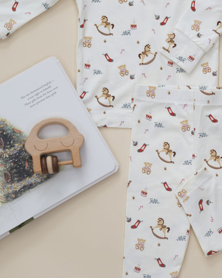 A flat-lay of a baby's long-sleeve pajama set featuring a white fabric with a holiday-themed print of rocking horses, teddy bears, candy canes, and red socks. The pajamas are styled alongside a wooden horse teether and an open children's book on a light beige background.