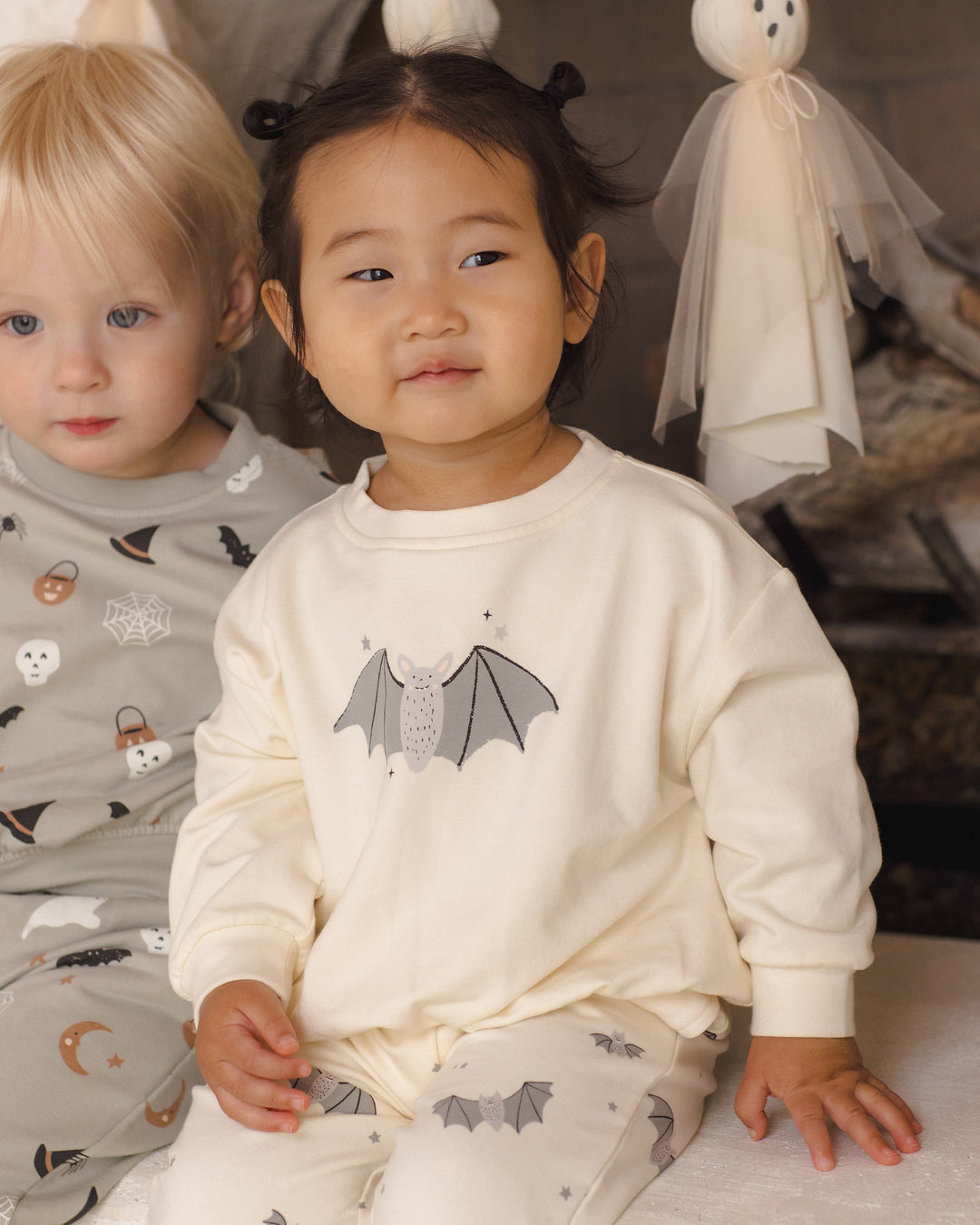 A toddler wearing a cream long-sleeve pajama top with a gray bat graphic and matching patterned pajama pants, seated indoors next to another child in a gray patterned pajama set.