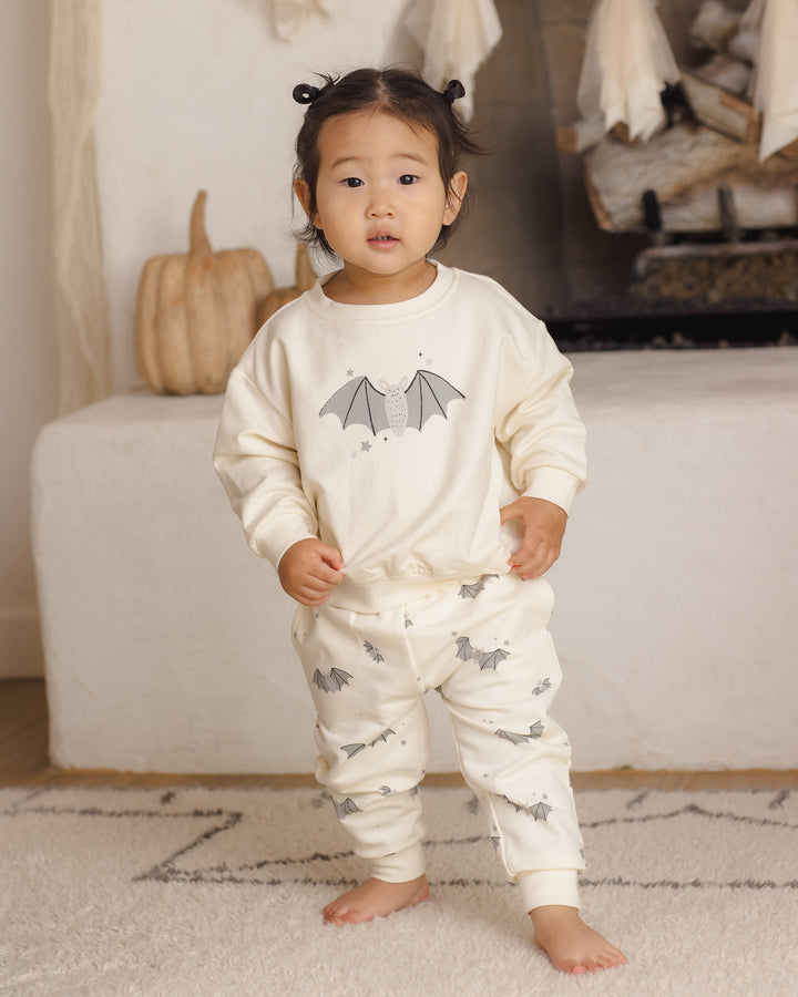 A toddler stands barefoot indoors wearing a cream-colored long-sleeve sweatshirt and matching jogger pants with gray bat prints. The background shows a cozy room setting with a fireplace, a small pumpkin, and a rug.