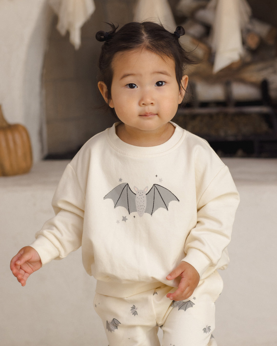 A toddler girl wearing a cream-colored pajama set with a bat graphic on the sweatshirt and matching bat prints on the pants, standing in a softly lit indoor setting.