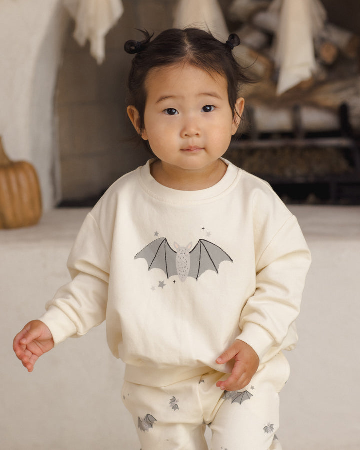 A toddler girl wearing a cream-colored pajama set with a bat graphic on the sweatshirt and matching bat prints on the pants, standing in a softly lit indoor setting.