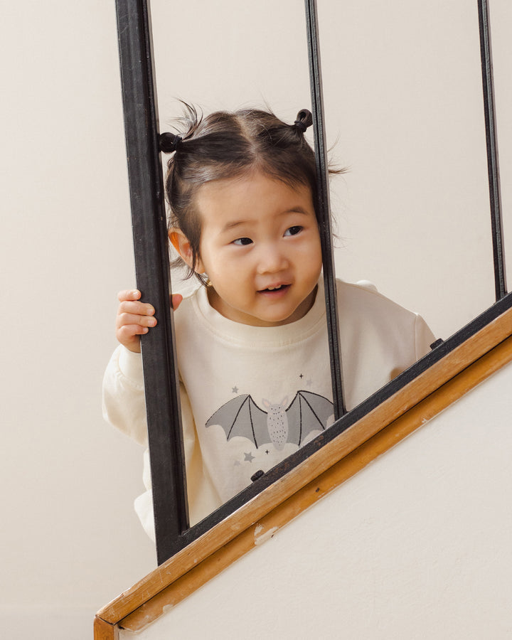 A young child with dark hair in two buns peeks over a black metal railing on a staircase, wearing a cream sweatshirt with a gray bat graphic.