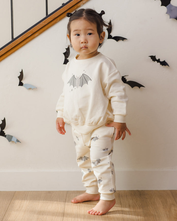 A toddler stands barefoot indoors wearing a cream-colored pajama set with a bat print. The set includes a long-sleeve sweatshirt and matching pants.