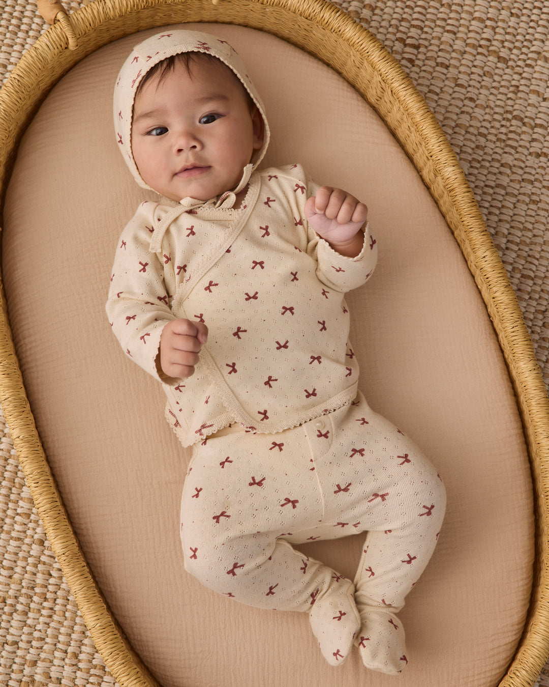A baby wearing a cream-colored footed sleeper and matching bonnet with a small red bow pattern, lying on a beige cushion inside a woven oval basket.