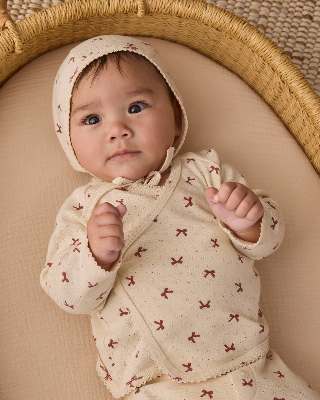 A baby lying on a beige cushion inside a woven bassinet, wearing a cream-colored hooded romper with a small red bow pattern.
