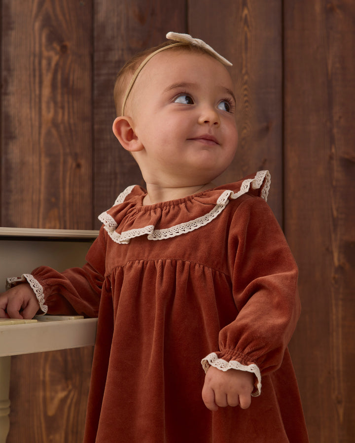 A baby girl wearing a rust-red velour dress with white lace trim and a matching cream bow headband, standing in front of a dark wood plank wall.