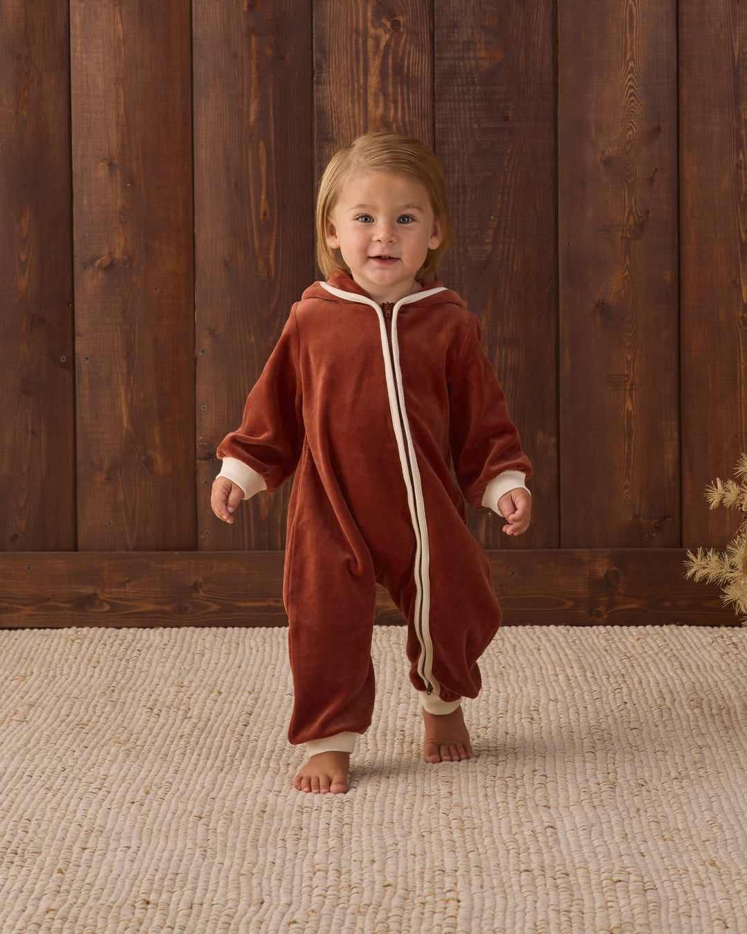 A toddler walking barefoot indoors wearing a rust-red hooded one-piece jumpsuit with cream trim and a front zipper. The child stands on a light beige woven rug in front of a dark wood plank wall.