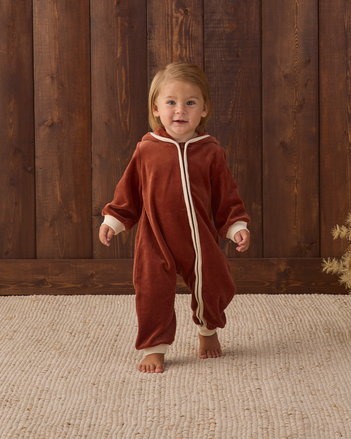 A toddler walking barefoot indoors wearing a rust-red hooded one-piece jumpsuit with cream trim and a front zipper. The child stands on a light beige woven rug in front of a dark wood plank wall.