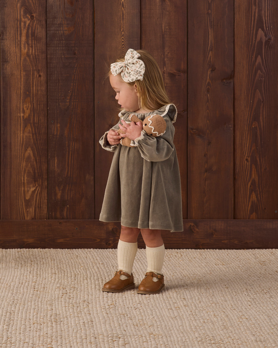 A toddler girl stands in profile against a dark wood plank wall, wearing a muted green velour dress with cream lace trim, a large cream bow headband, cream knee-high socks, and brown moccasin-style shoes.