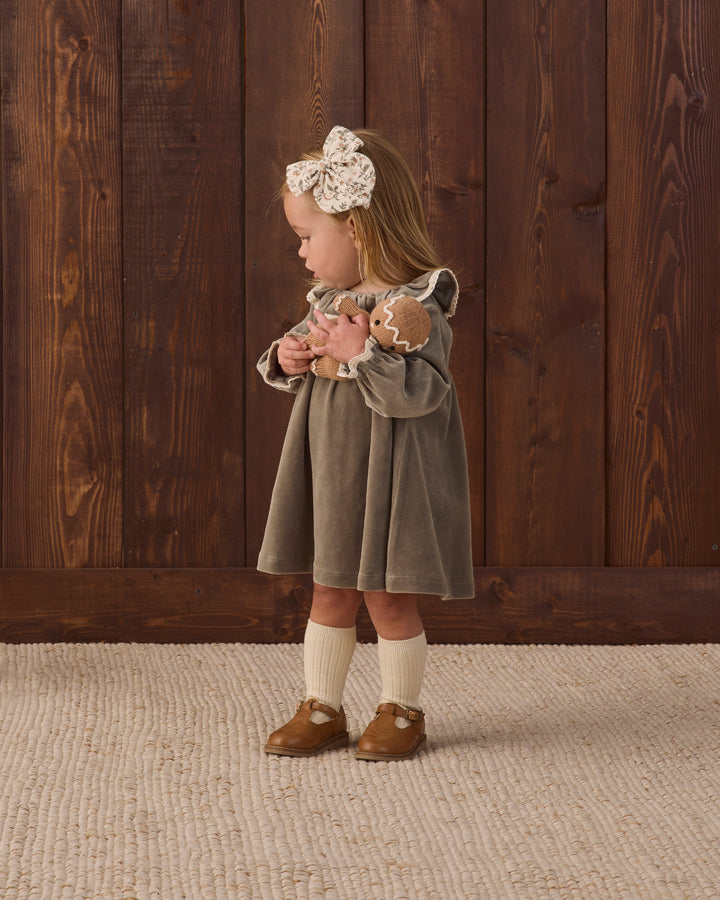 A toddler girl stands in profile against a dark wood plank wall, wearing a muted green velour dress with cream lace trim, a large cream bow headband, cream knee-high socks, and brown moccasin-style shoes.