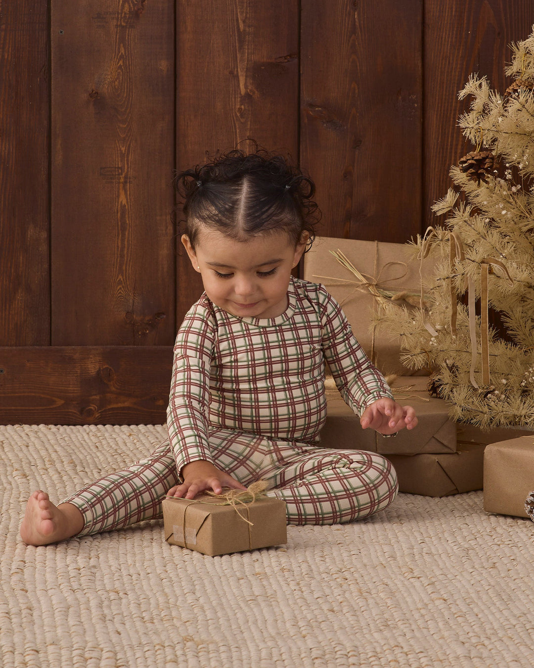 A toddler sits on a beige woven rug in front of a dark wood plank wall, wearing a long-sleeve plaid pajama set in muted red, green, and beige tones. The child is holding a small wrapped gift box, with wrapped presents and a decorative tree in the background.