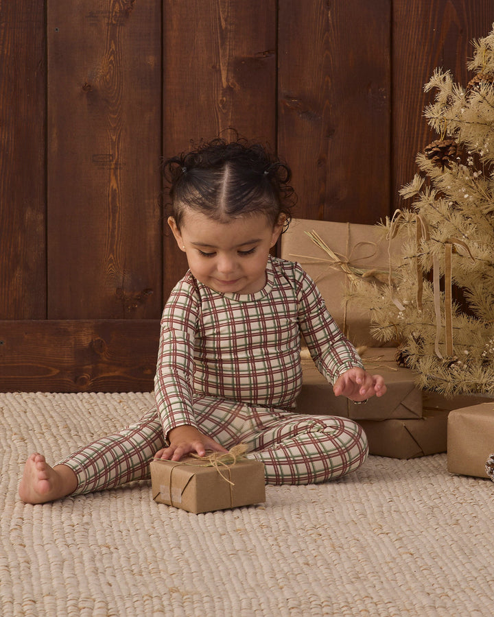 A toddler sits on a beige woven rug in front of a dark wood plank wall, wearing a long-sleeve plaid pajama set in muted red, green, and beige tones. The child is holding a small wrapped gift box, with wrapped presents and a decorative tree in the background.
