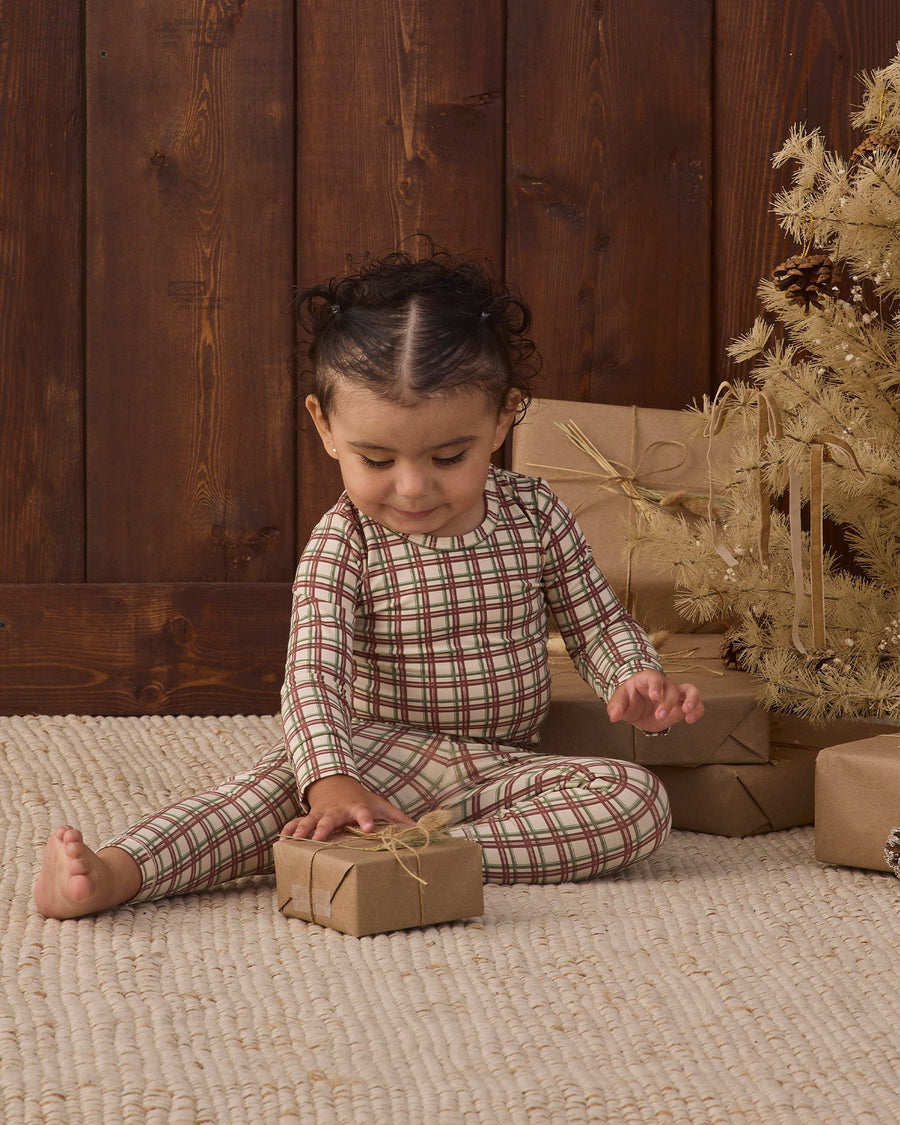 A toddler sits on a beige woven rug in front of a dark wood plank wall, wearing a long-sleeve plaid pajama set in muted red, green, and beige tones. The child is holding a small wrapped gift box, with wrapped presents and a decorative tree in the background.
