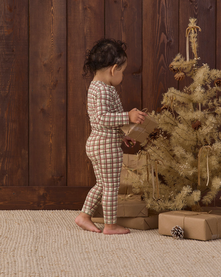 A toddler stands barefoot in front of a rustic dark wood wall, wearing a long-sleeve plaid pajama set in beige, brown, and muted red. The child is holding a wrapped gift next to a decorated Christmas tree with wrapped presents on the floor.