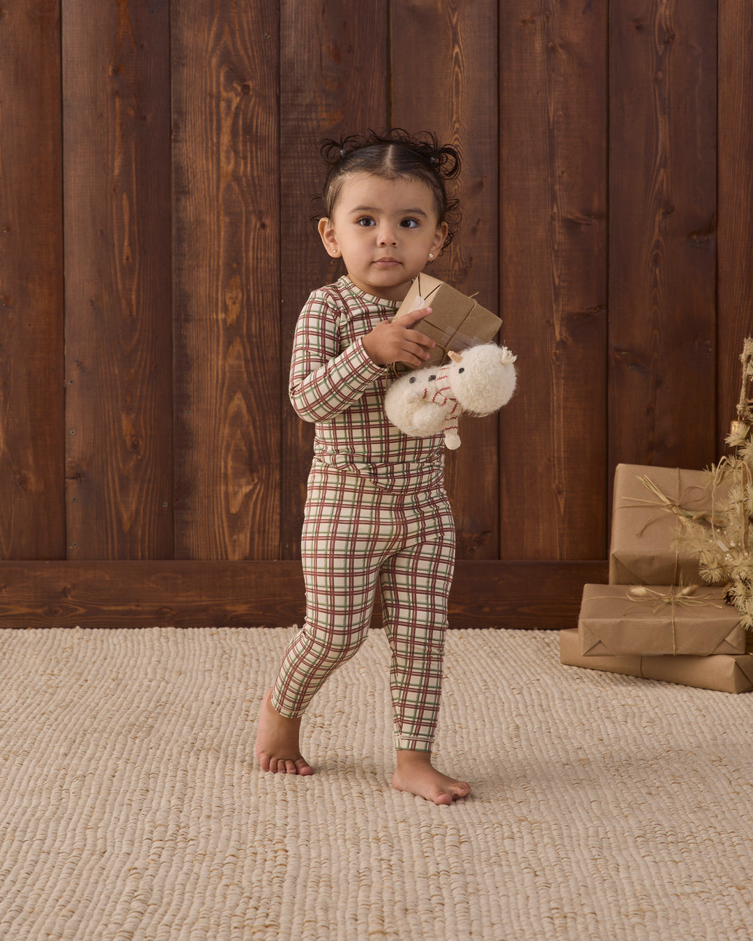 A toddler stands barefoot on a textured beige rug in front of a dark wood plank wall, wearing a brown and beige plaid long-sleeve pajama set and holding a small brown gift box with a white plush toy.