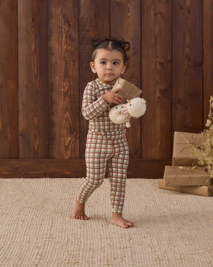A toddler stands barefoot on a textured beige rug in front of a dark wood plank wall, wearing a brown and beige plaid long-sleeve pajama set and holding a small brown gift box with a white plush toy.