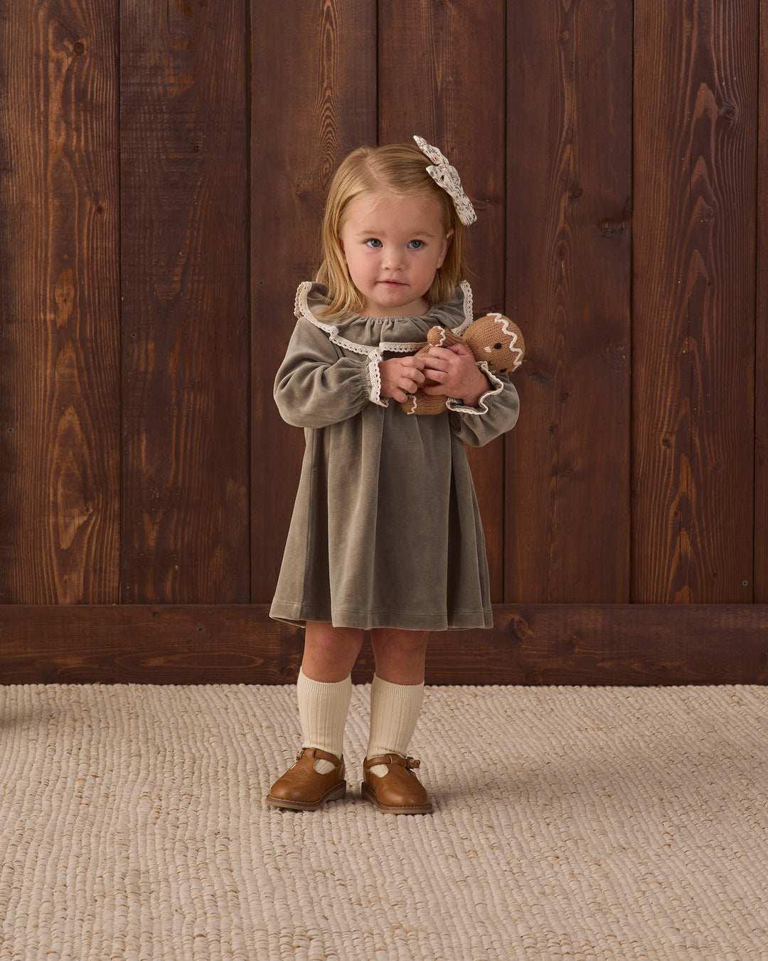 A young girl stands against a dark wood plank wall wearing a muted olive green dress with cream lace trim, cream knee-high socks, and brown moccasin-style shoes. She holds a small stuffed animal and wears a cream bow in her hair.