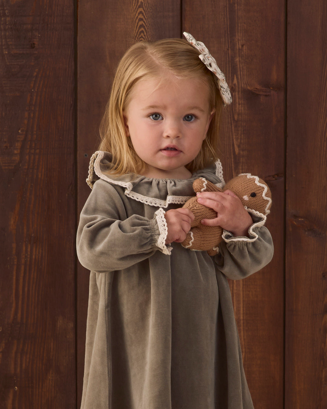 A young girl with blonde hair wearing a olive green long-sleeve velvet dress with cream lace trim and a matching hair bow, holding a small knitted stuffed animal against a dark wood plank background.