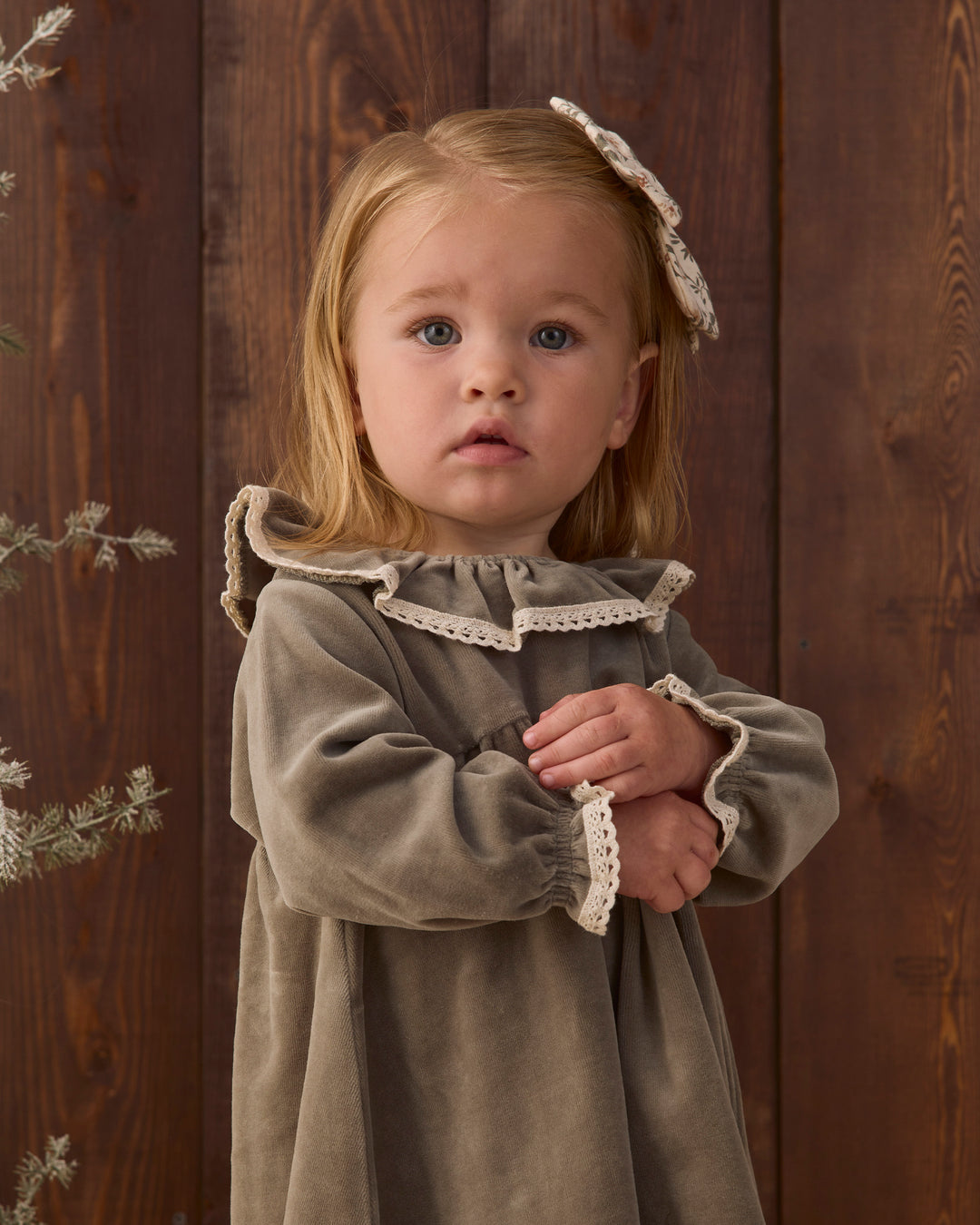 A young girl with blonde hair wearing a olive green velvet long-sleeve dress with cream lace trim and a matching lace hair bow, standing against a dark wooden plank background.