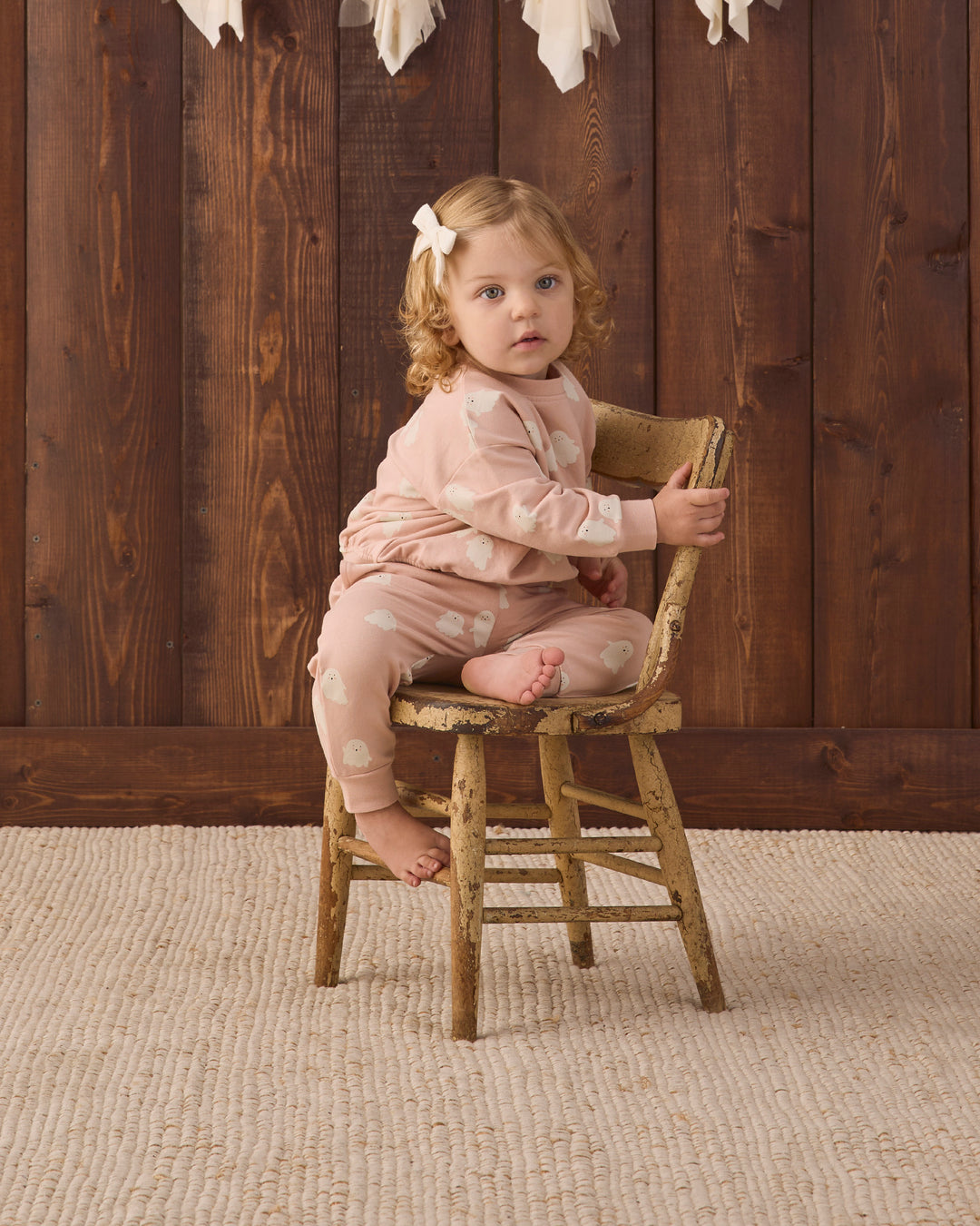 A toddler girl with curly light brown hair wearing a light pink sweat set with white ghosts sits on a rustic wooden stool in front of a dark wood plank wall.