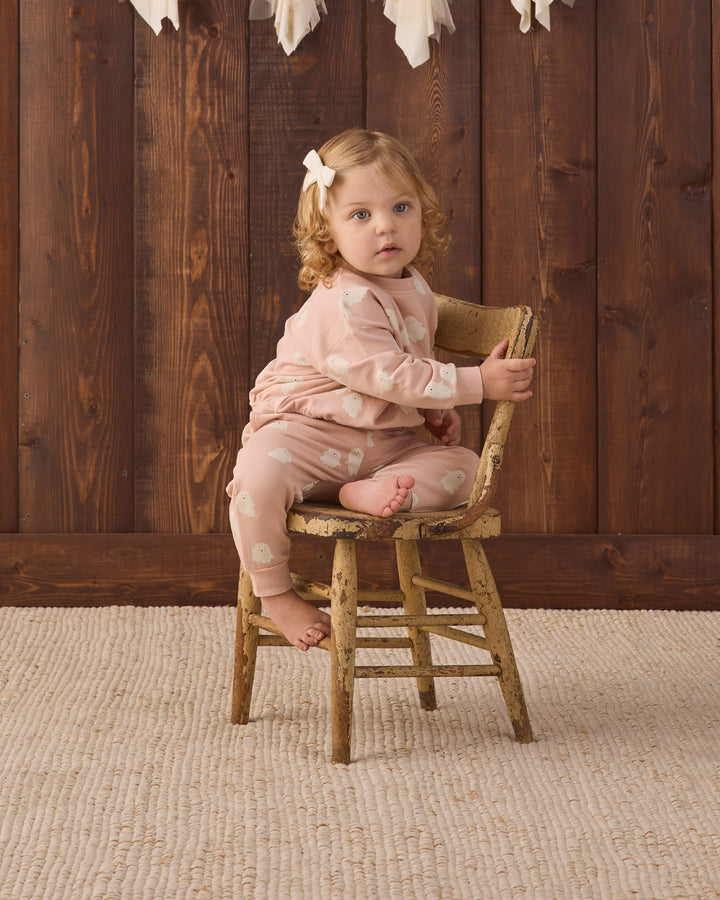A toddler girl with curly light brown hair wearing a light pink sweat set with white ghosts sits on a rustic wooden stool in front of a dark wood plank wall.