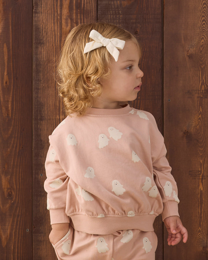 A young child with blonde curly hair wearing a light pink sweatshirt and matching pants with white ghost prints, standing against a dark wooden plank background.