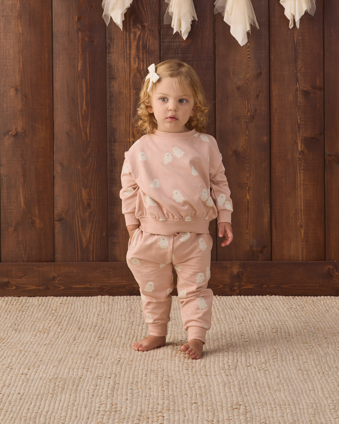A toddler girl stands barefoot on a textured beige rug in front of a dark wood plank wall, wearing a matching dusty pink two-piece sweat set with white ghosts. The set includes a long-sleeve top and matching pants.