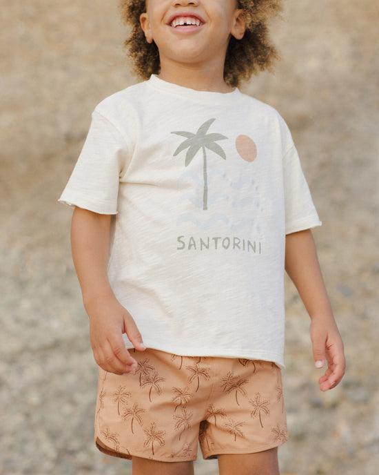 boy modeling the raw edge tee with the word 'santorini' written across the chest, a palm tree, sun and waves. 