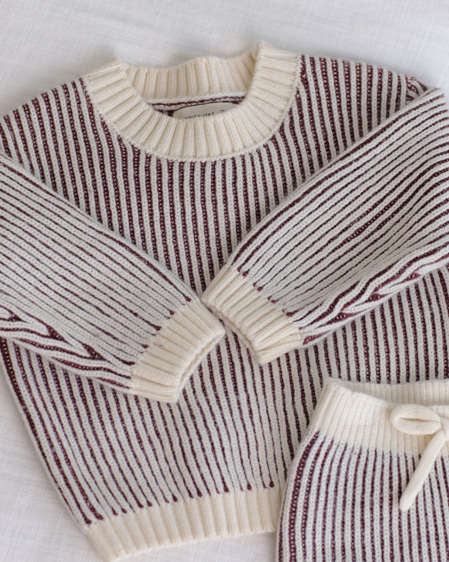 A close-up of a ribbed knit sweater laid flat on a white textured fabric background. The sweater has vertical dark burgundy stripes on an off-white base, with cream-colored ribbed trim at the crew neck, cuffs, and hem.