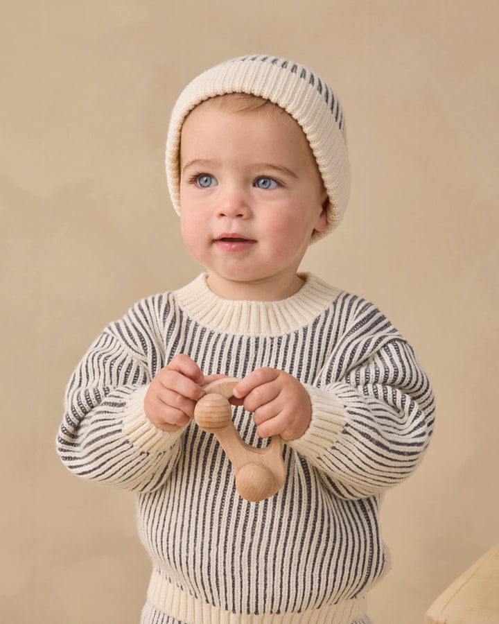 A baby wearing a cream knit beanie and a cream sweater with thin dark gray stripes, holding a wooden teething toy against a warm beige background.