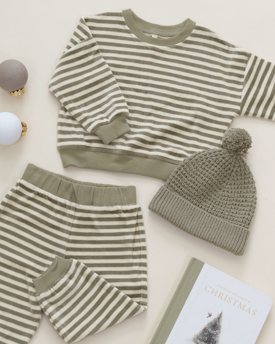A baby holiday pajama set featuring a long-sleeve top and matching pants in a beige and sage green stripe pattern, styled with a matching knit pom-pom beanie, decorative ornaments, and a book on a light beige background.