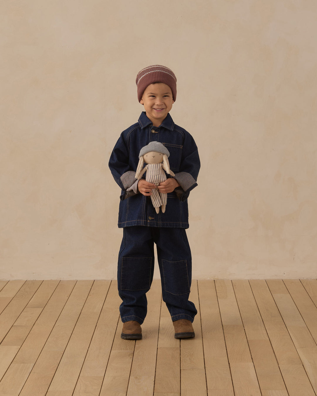 A young child stands on a light wood floor against a warm beige wall, wearing a dark blue denim jacket and matching denim pants, with a striped knit beanie and holding a small stuffed doll.