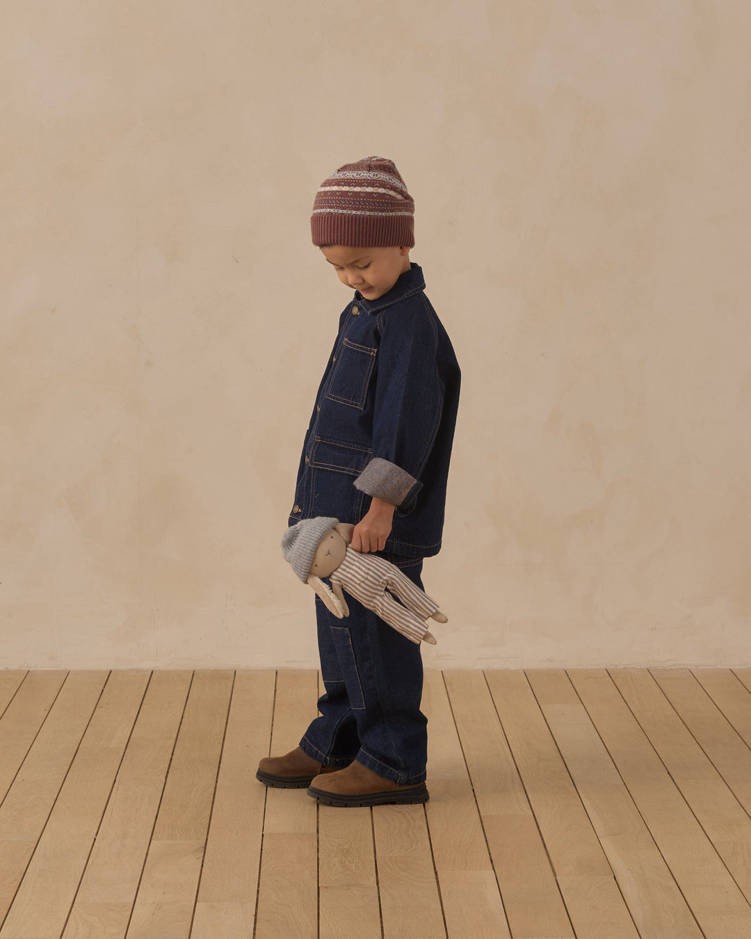 A toddler stands in profile wearing a dark blue denim work jacket and matching denim pants, with a burgundy and cream striped knit beanie and brown boots. The child is holding a gray and white striped plush bunny against a warm beige textured wall and light wood plank floor.