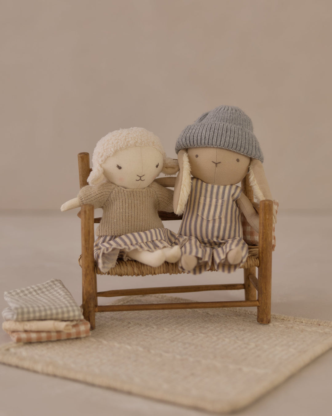 Two soft fabric dolls sitting on a small wooden chair, styled with neutral-toned textiles against a warm beige background.