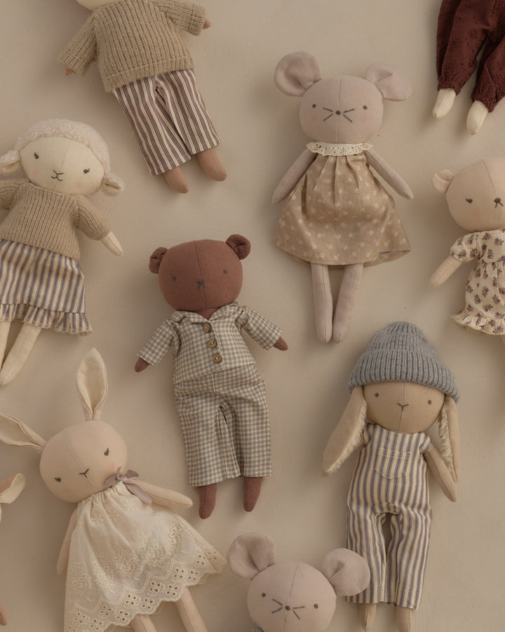 A collection of handmade fabric dolls and stuffed animals in soft neutral tones, including a teddy bear in a gingham outfit, a rabbit in a lace dress, a bunny in striped overalls with a knit blue hat, and other plush toys arranged on a light beige background.