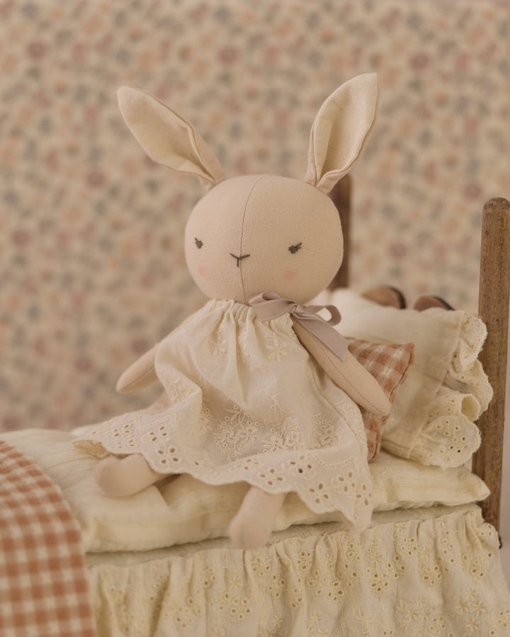 A soft plush bunny doll with long ears sits on a miniature bed dressed in a cream lace dress, set against a vintage floral wallpaper background.