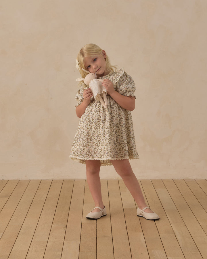 A young blonde girl stands on a light wood floor against a textured beige wall, wearing a cream and brown floral print dress and holding a small white stuffed animal.