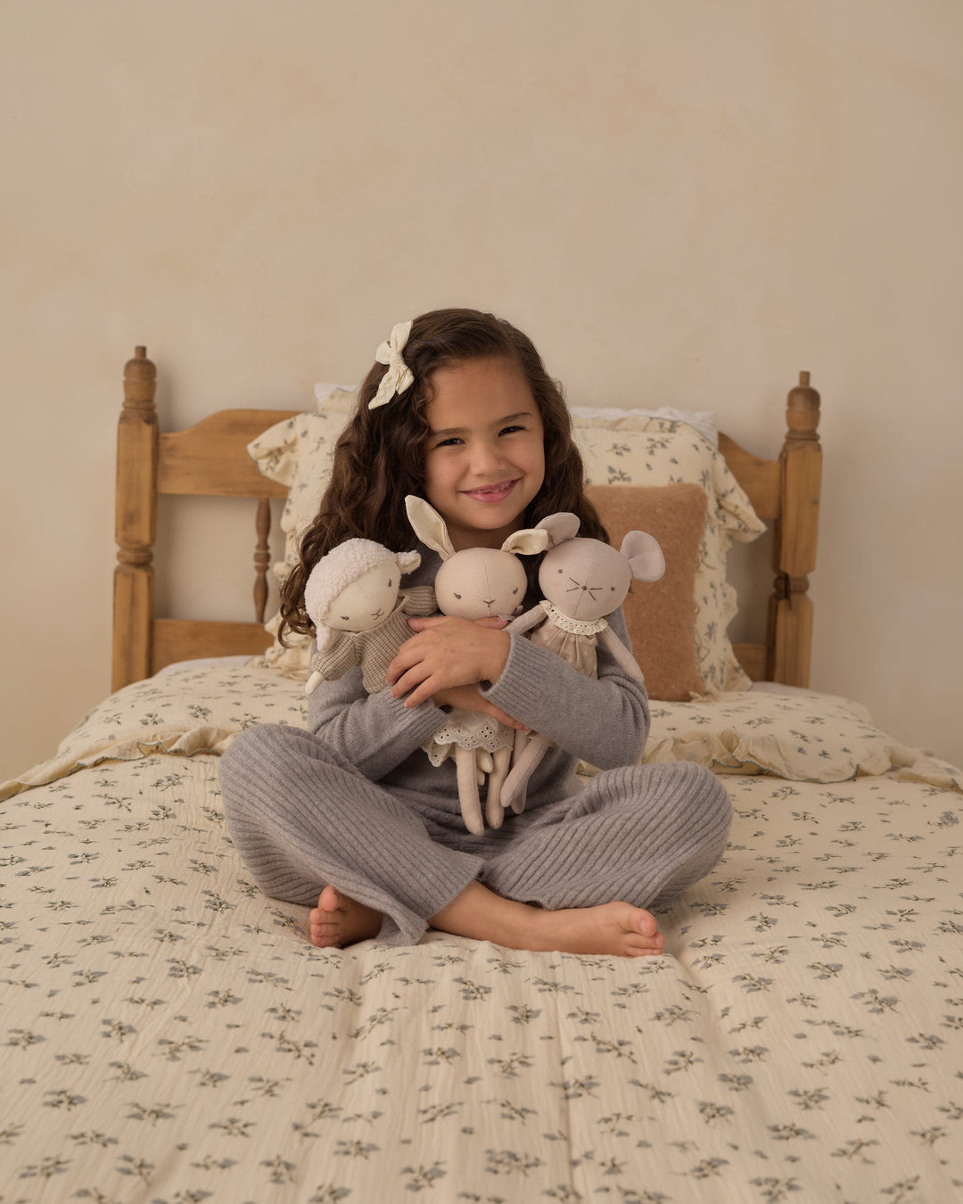 A young girl with long brown hair sits cross-legged on a floral bed, holding three small plush stuffed animals. She is wearing a gray ribbed pajama set, with a wooden bed frame and cream-colored bedding in the background.