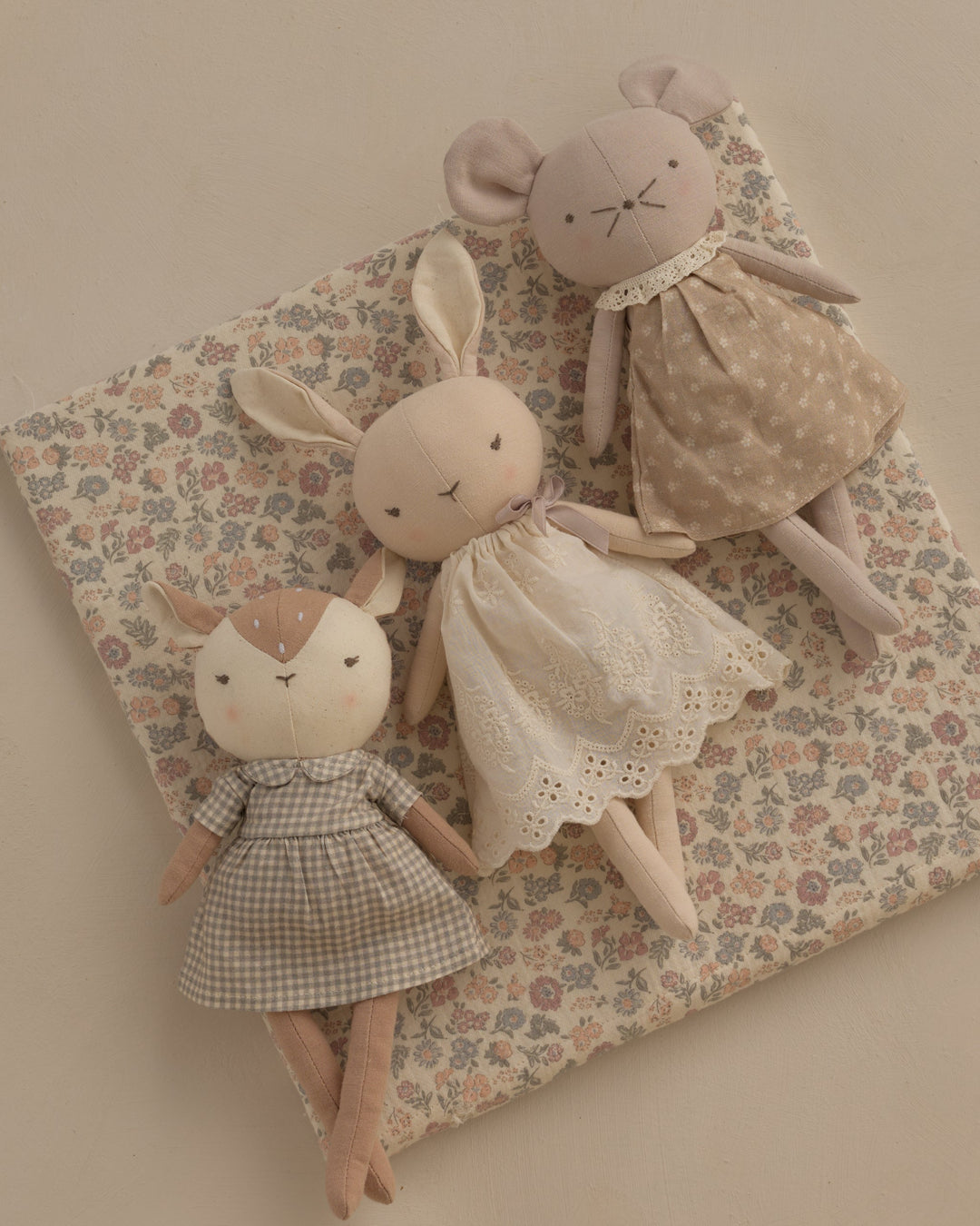 Three handmade plush bunny dolls in soft neutral tones, each wearing a different embroidered dress, arranged on a floral cushion against a warm beige background.